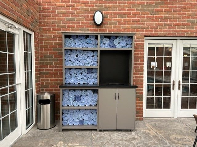 Outdoor cabinet with rolled blue towels, cabinet doors, and a trash can, set against a brick wall with glass doors.