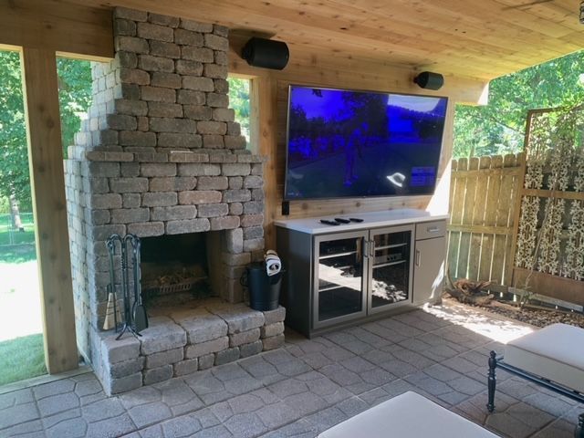 Outdoor patio with stone fireplace, TV, and speakers. Wooden ceiling and cabinet.