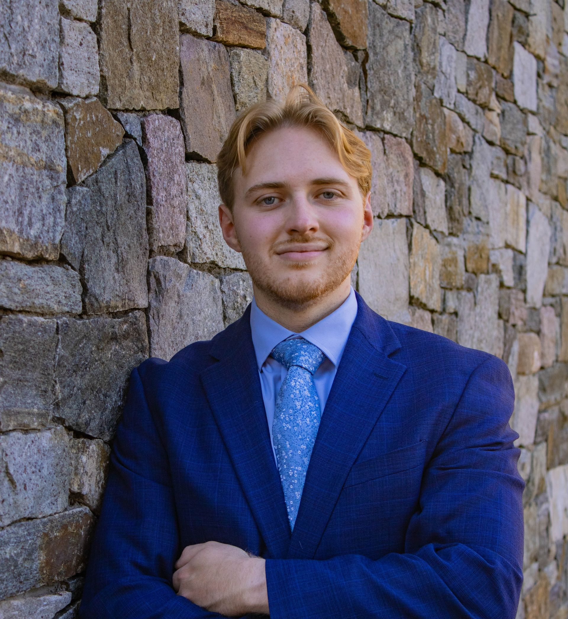 Young man with blond hair in a blue suit smiles, arms crossed, leaning against a stone wall.