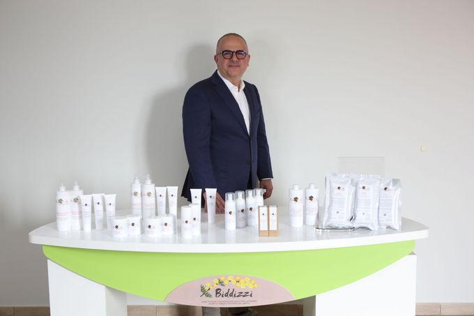 Un uomo in giacca e cravatta si trova dietro un bancone curvo bianco e verde che espone numerosi prodotti cosmetici bianchi.
