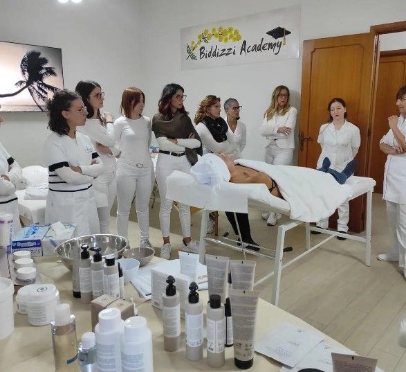 Presso l'Accademia Biddizzi, alcuni studenti in uniforme bianca osservano una sessione di formazione professionale attorno a un lettino da massaggio.