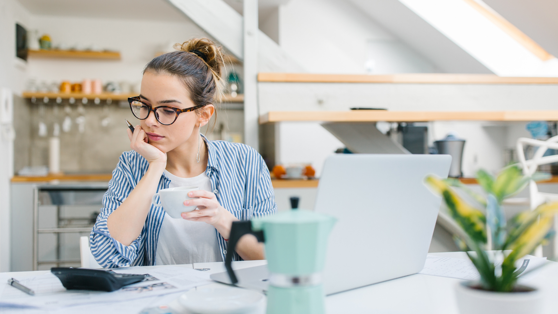 Woman in glasses at a desk, holding a mug and looking at a laptop in a bright home office.
