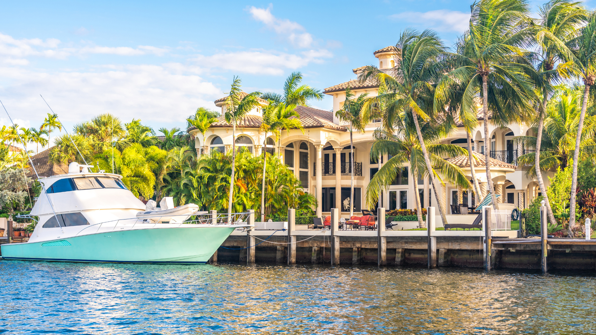 Luxury yacht docked beside a waterfront mansion with palm trees under a bright blue sky