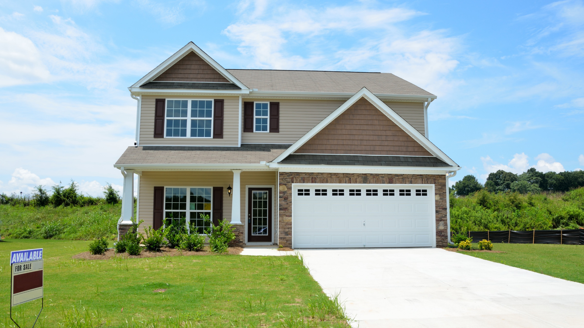Two-story tan house with a white garage door and driveway on a green lawn under a blue sky