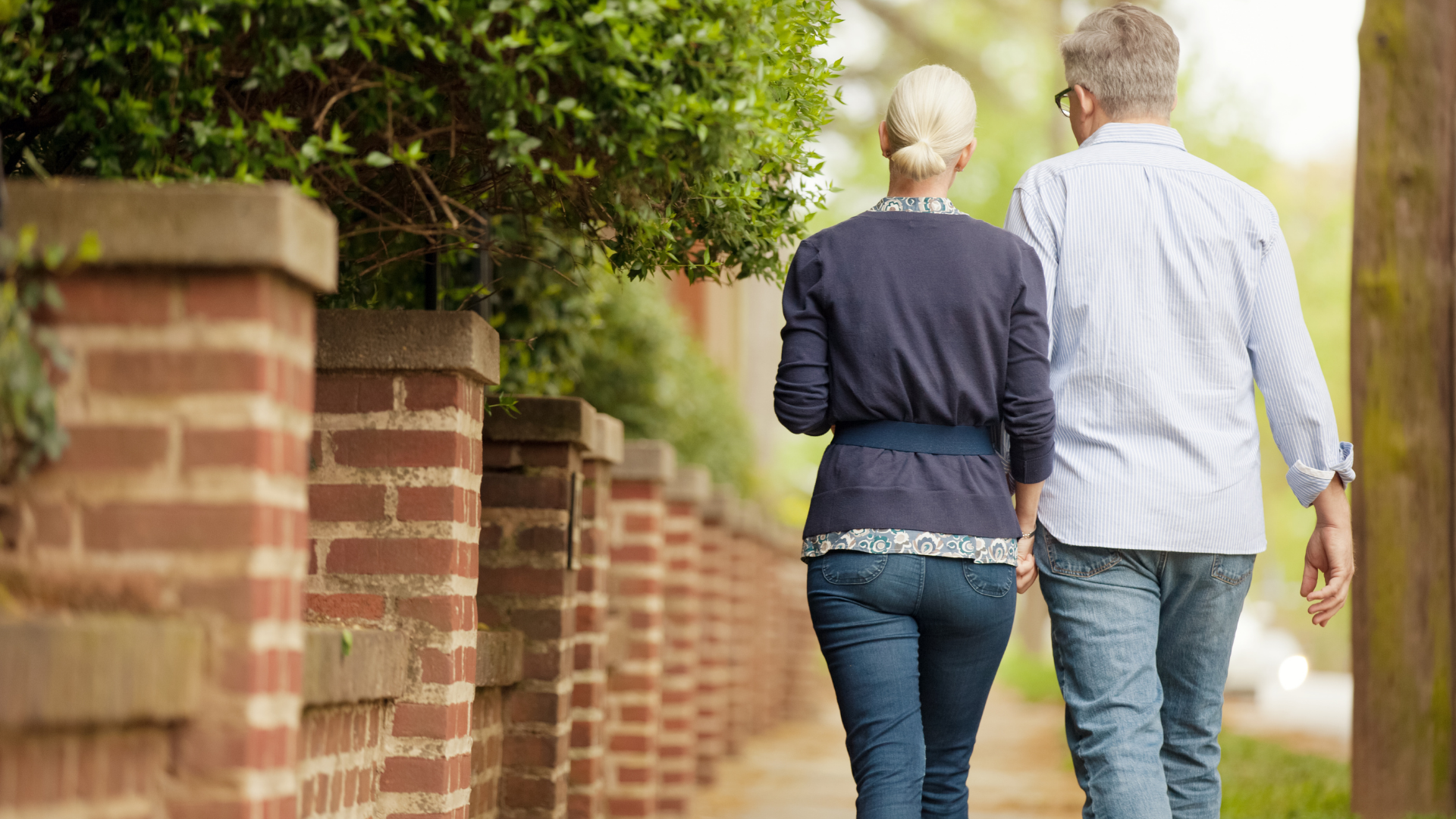 Two people walking away along a tree-lined brick path beside a red brick wall.