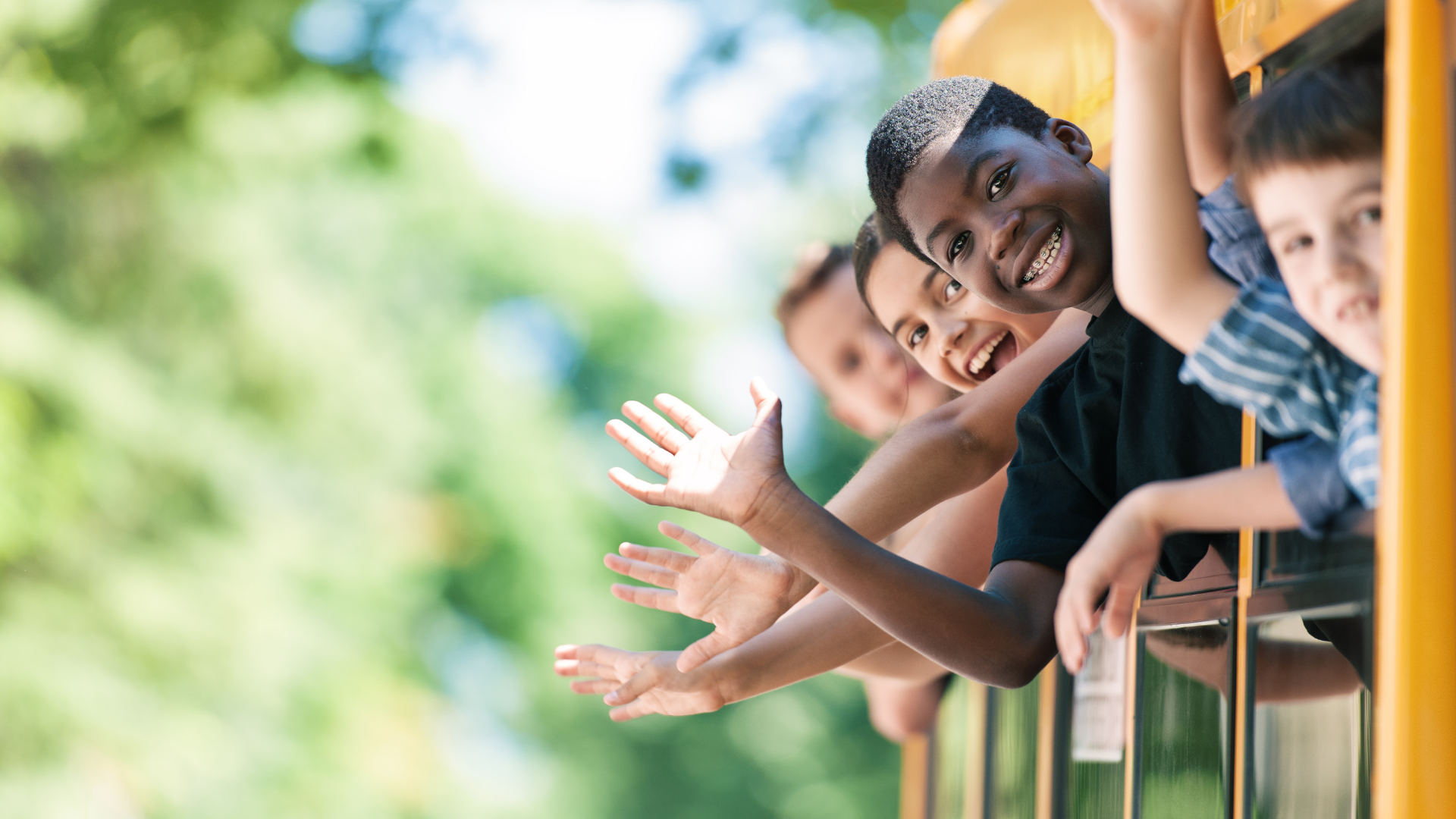 Children leaning over a railing, smiling and waving outdoors in bright sunlight.