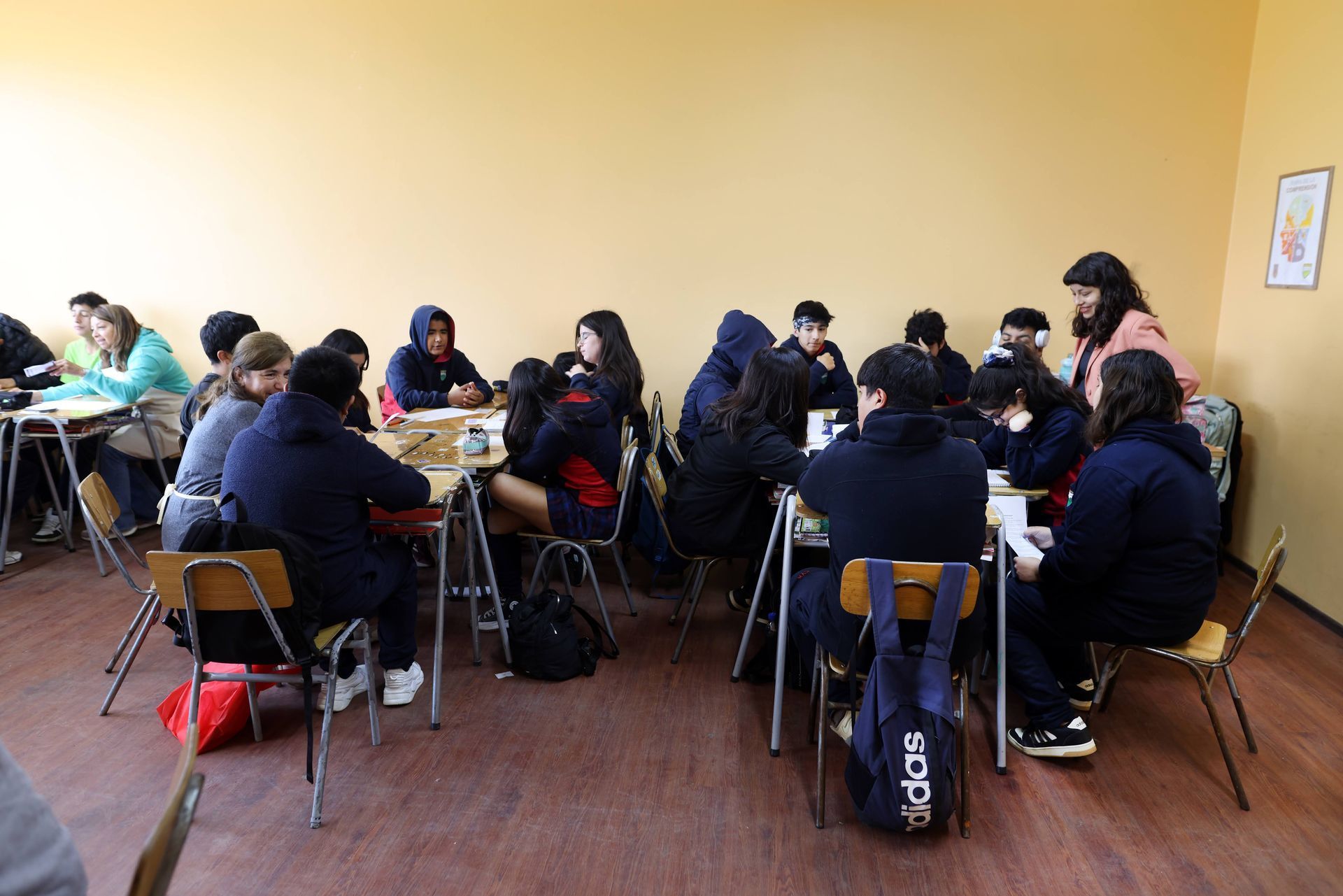 Estudiantes sentados en mesas en un aula; algunos comiendo, otros trabajando. Paredes amarillas, suelo de madera.