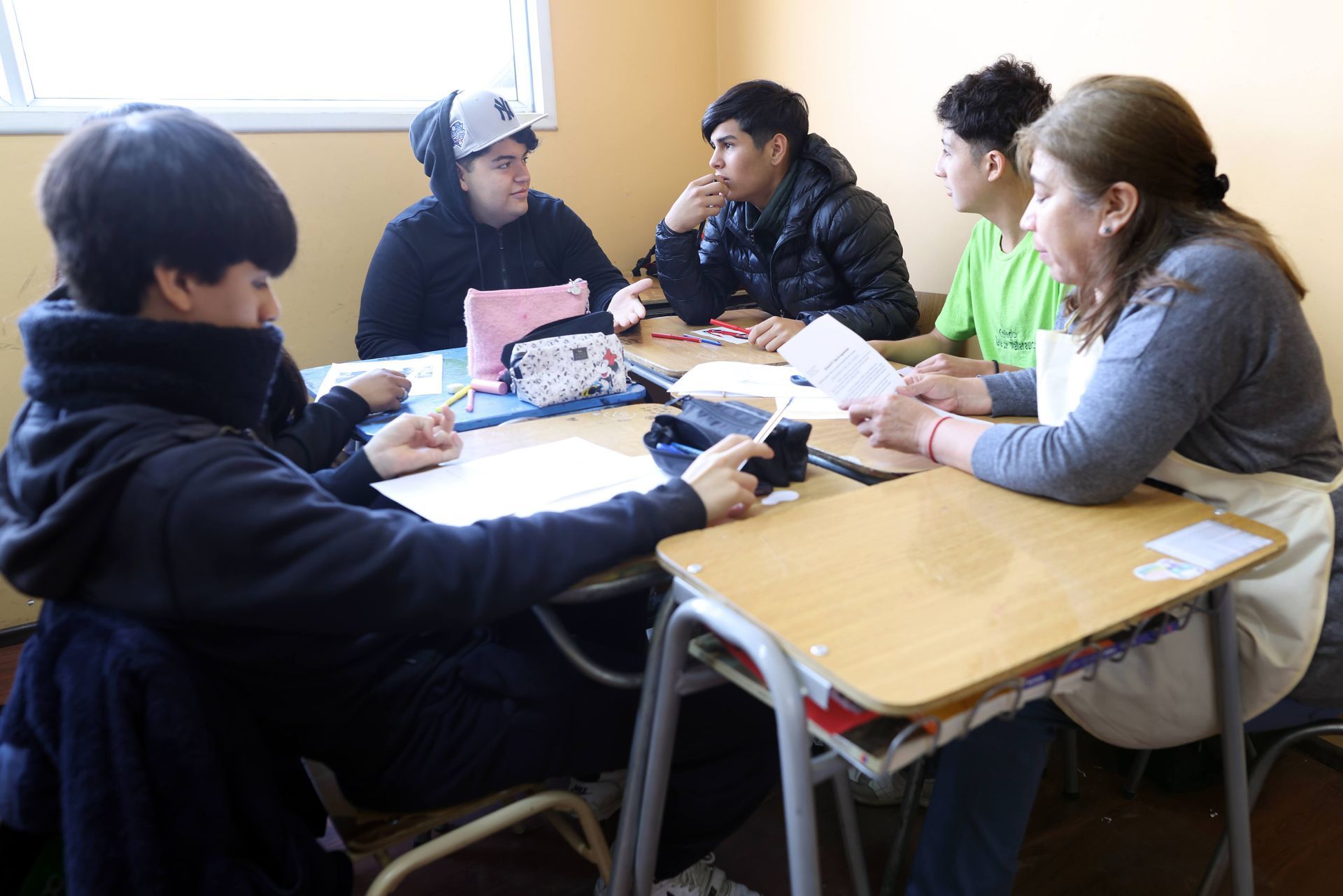 Estudiantes y profesor en un aula, reunidos alrededor de un escritorio, mirando papeles y conversando.