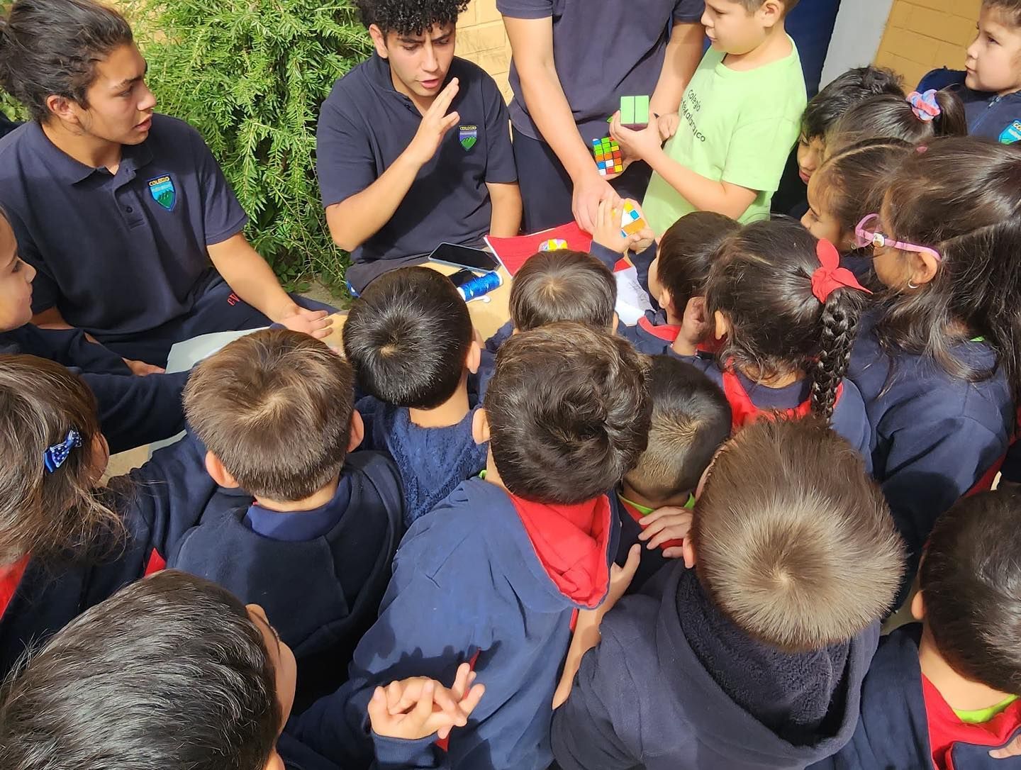 Los estudiantes se reúnen alrededor de una mesa al aire libre y observan una demostración que involucra objetos pequeños.