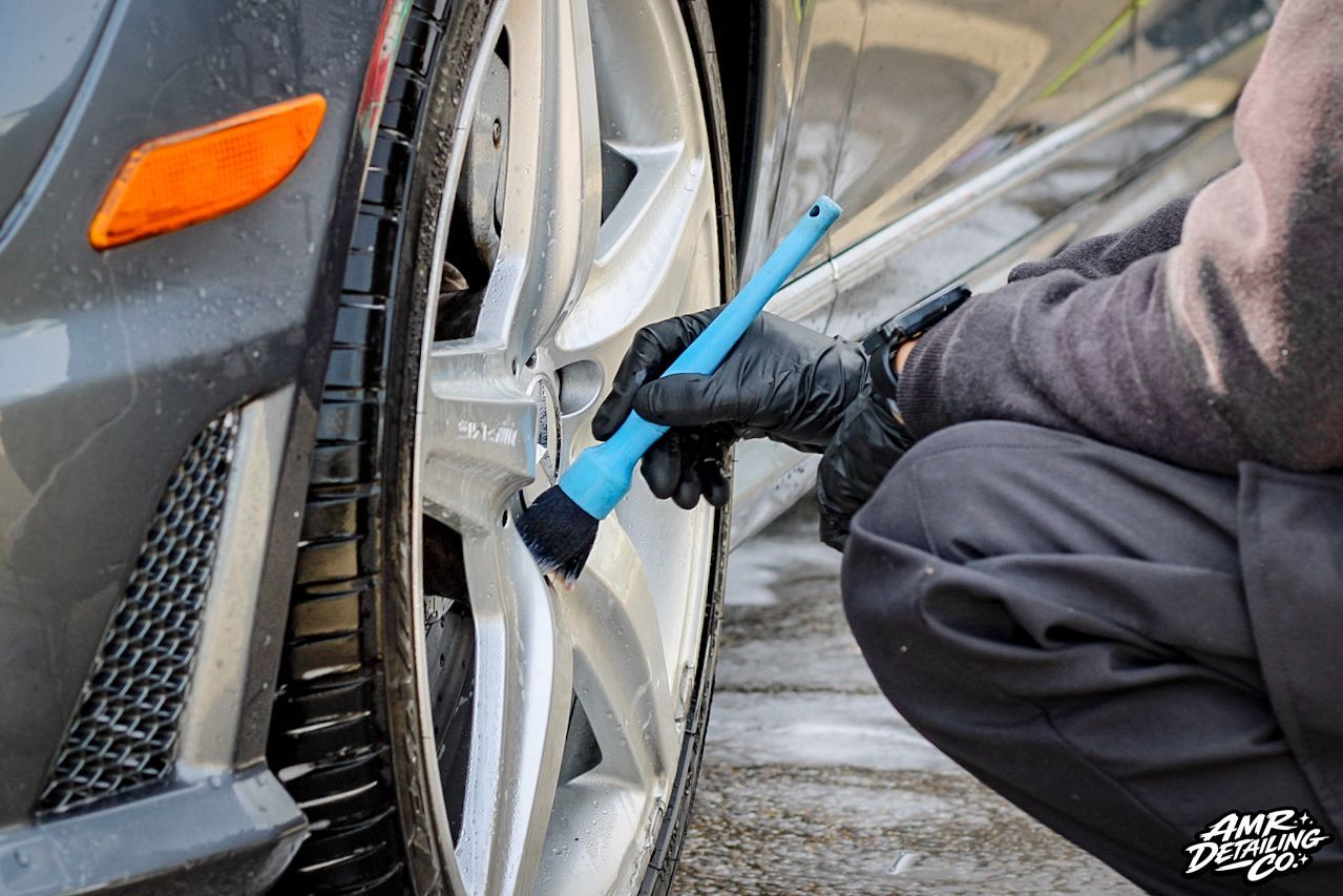 Gloved hand scrubs a car’s white wheel with a blue brush during a wash.