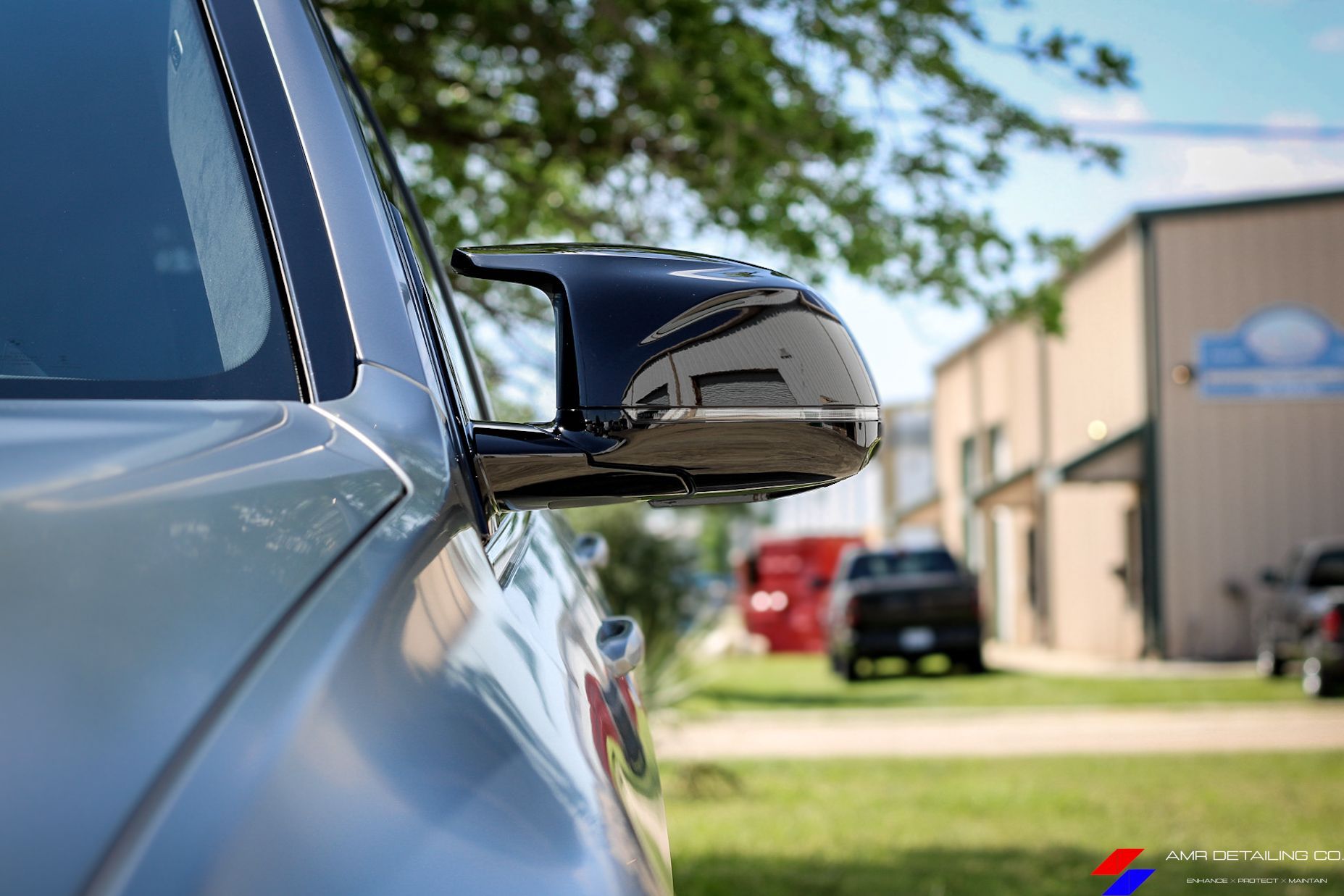 Close-up of a black car side mirror reflecting a building and trees on a sunny street.