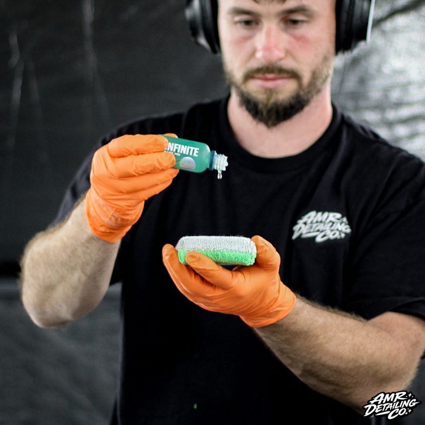 Gloved man applying green cleaning spray to a sponge against a dark background