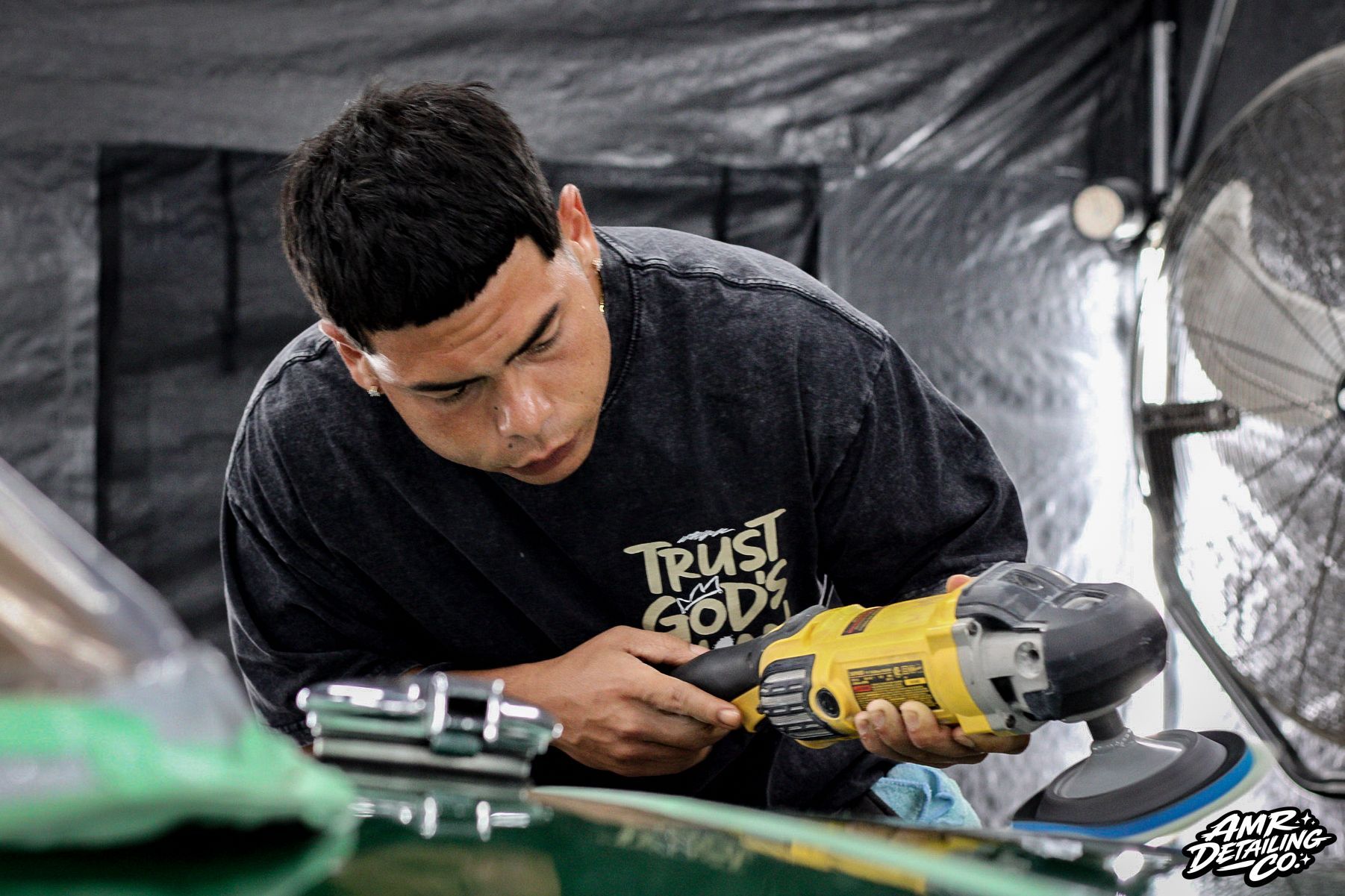 Man in black shirt using a yellow power sander on a green surface in a workshop