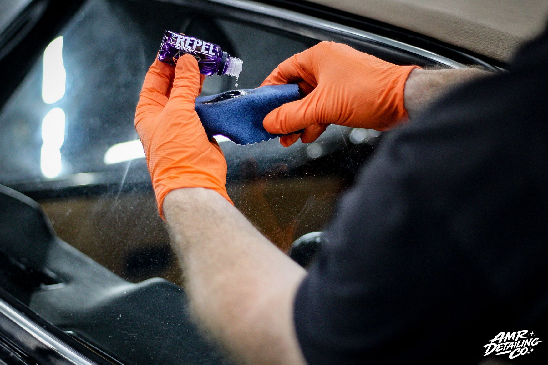 Orange-gloved hands apply a purple detailing product to a car windshield