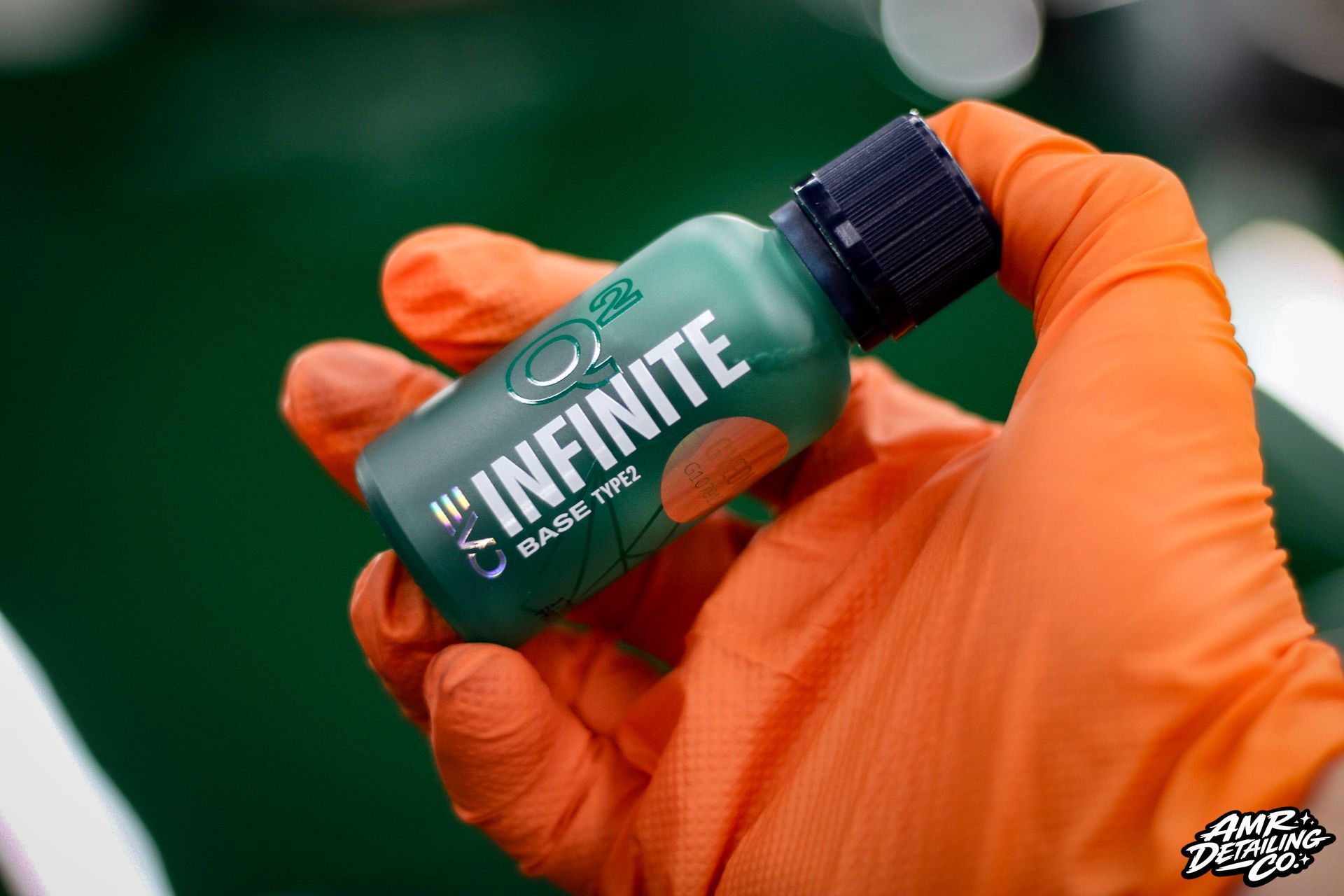 Orange-gloved hand holding a green Infinite bottle with a black cap against a blurred background