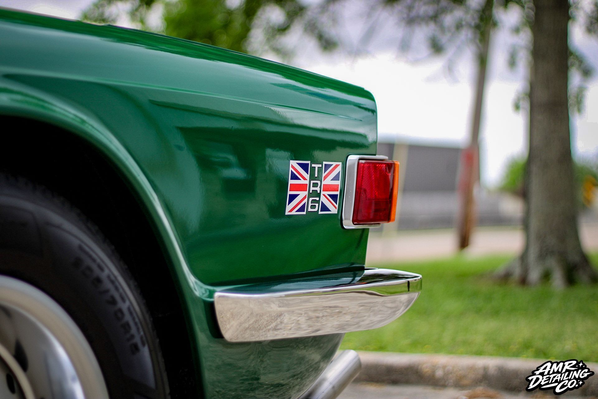 Close-up of a green classic car’s rear with a Union Jack badge and chrome bumper.
