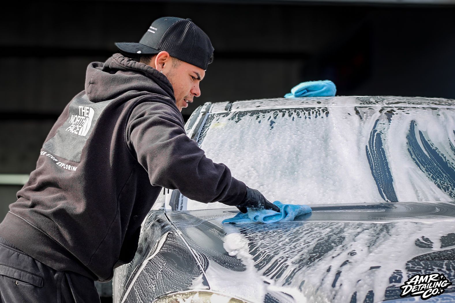 Man washing a car windshield with a blue sponge, covered in soap suds