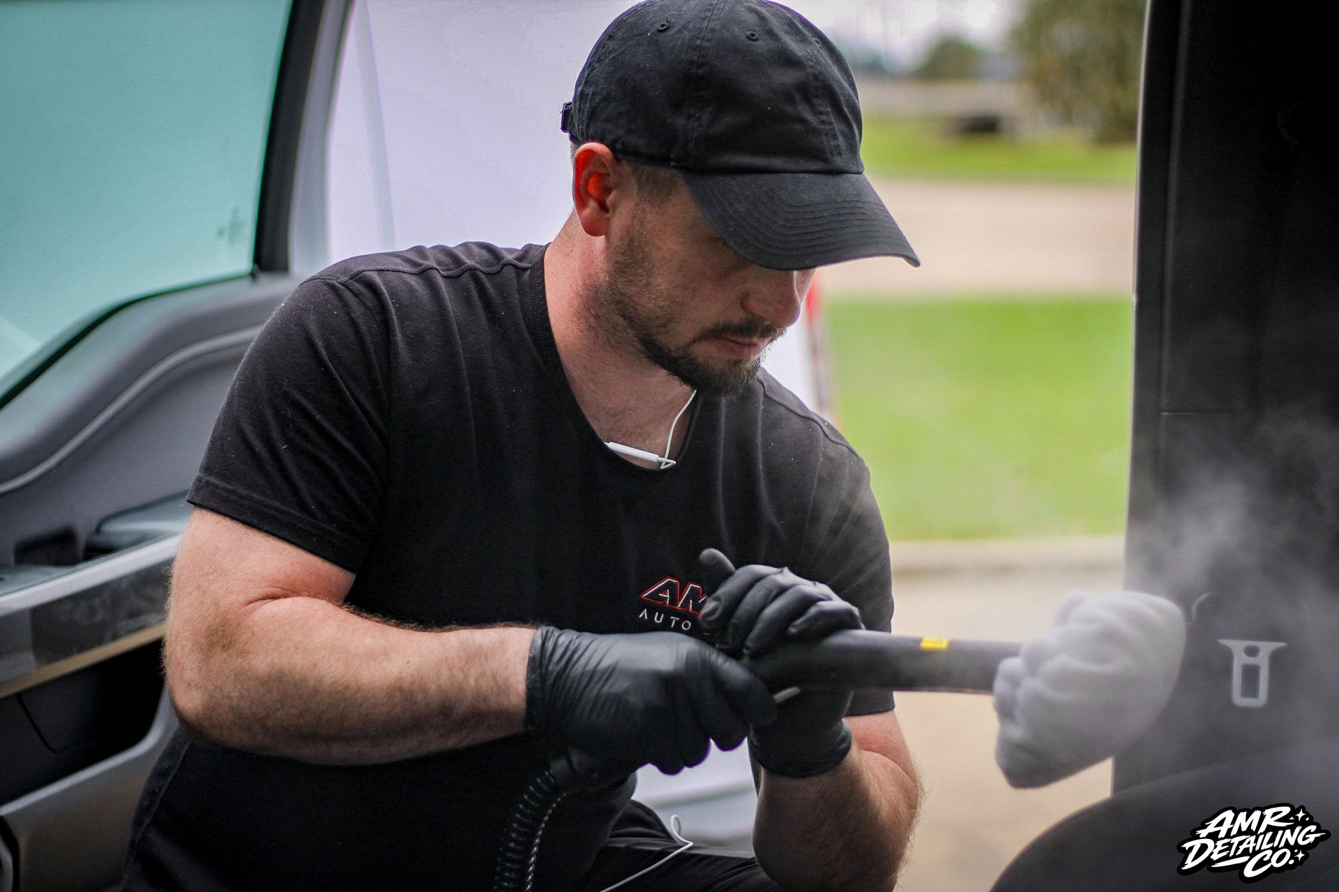 Technician in black gloves and cap using a tool near a car panel, with steam or smoke rising.