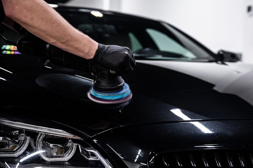 Gloved hand polishing a black car hood with a buffer tool