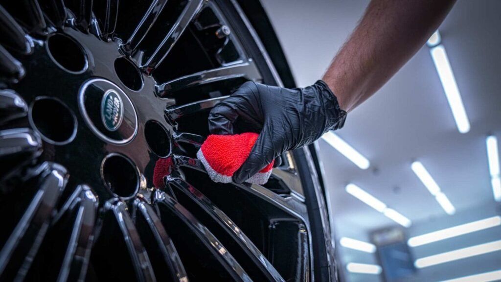 Gloved hand polishing a black alloy car wheel with a red cloth in a garage
