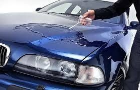 A person wiping a blue car’s hood with a cloth, showing a dented front end.