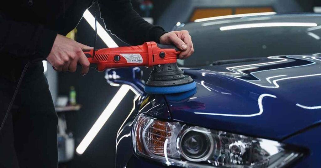 Person polishing a blue car hood with a red buffer in a garage