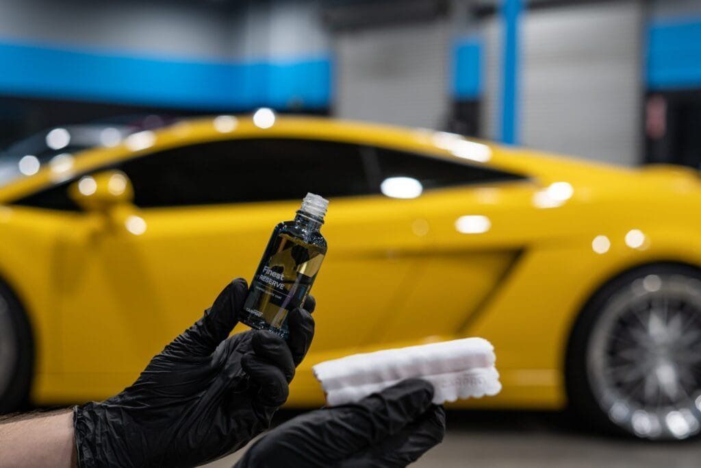 Gloved hands hold a car detailing spray bottle and applicator in front of a yellow sports car.
