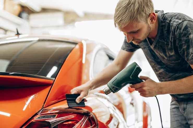Technician applying vinyl wrap to a red sports car with a heat gun in a workshop