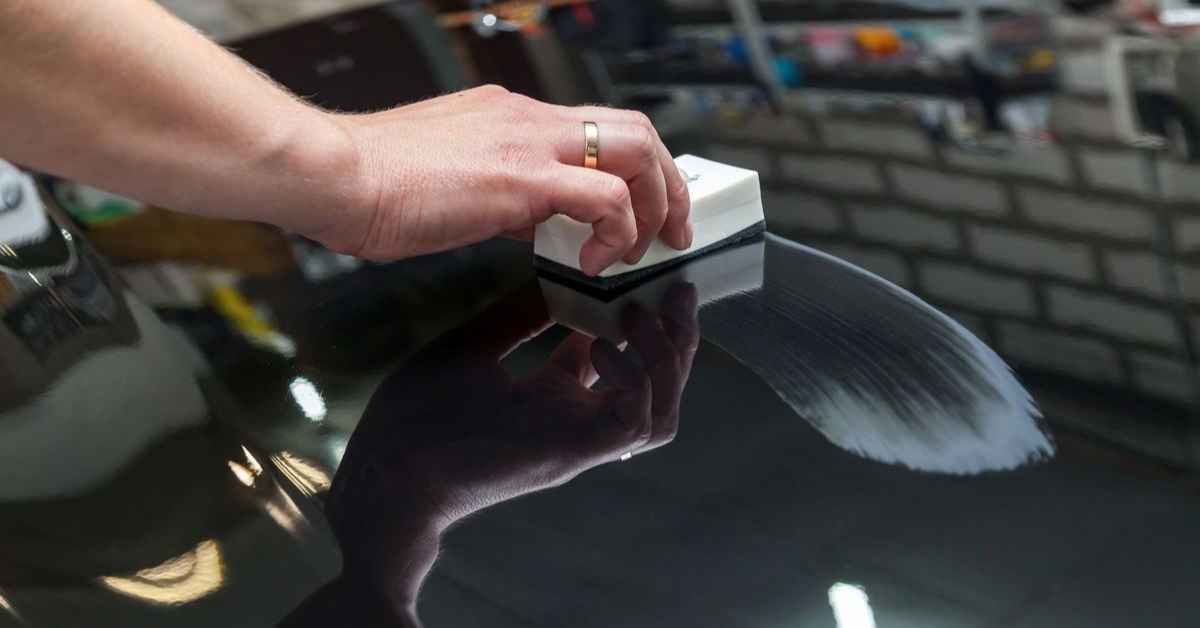Hand using a white block to polish a glossy black car hood outdoors