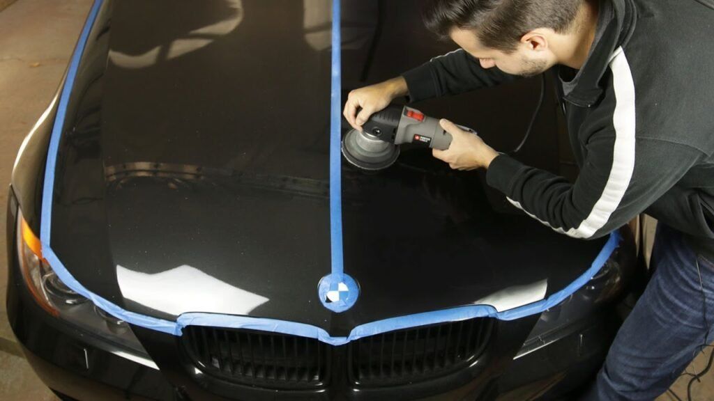 Man polishing the hood of a black car with a buffer, blue tape marking sections.