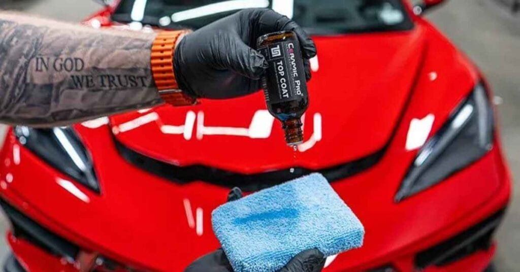 Gloved hand spraying car polish onto a red sports car’s hood with a blue microfiber cloth nearby