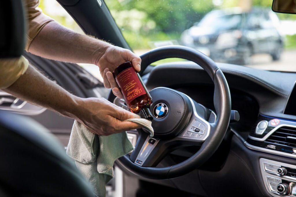 Person cleaning a BMW steering wheel with a spray bottle and cloth inside a car