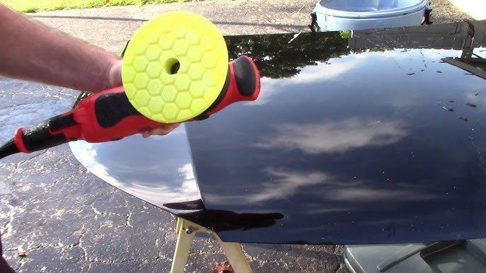 Hand holding a red-and-black buffer polishing a shiny black car hood with yellow pad