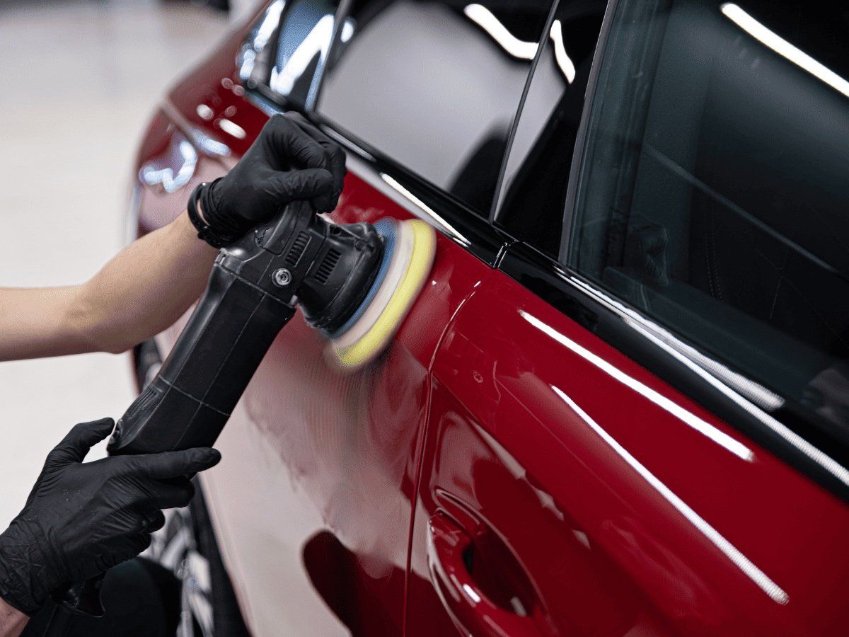 Gloved hands buffing a red car door with a power polisher in an auto shop