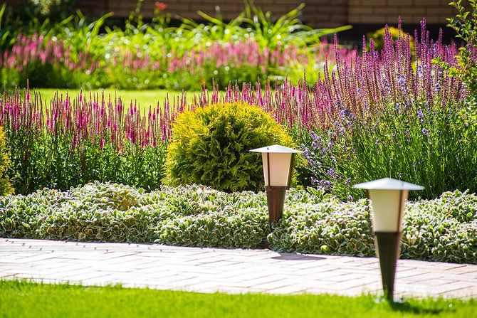 Colorful flower garden with pink blooms, lush greenery, and two pathway lights beside a wooden walkway