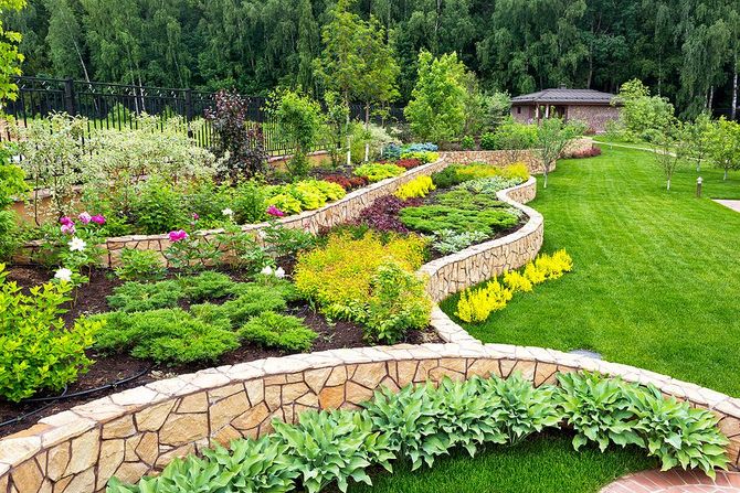 Curved stone garden terrace with colorful flowers, shrubs, and lush green lawn against a wooded backdrop