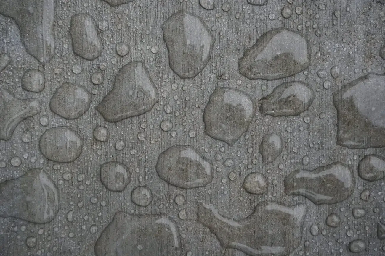 Water droplets on a gray, textured surface. Some droplets are large and irregular; others are small and circular.