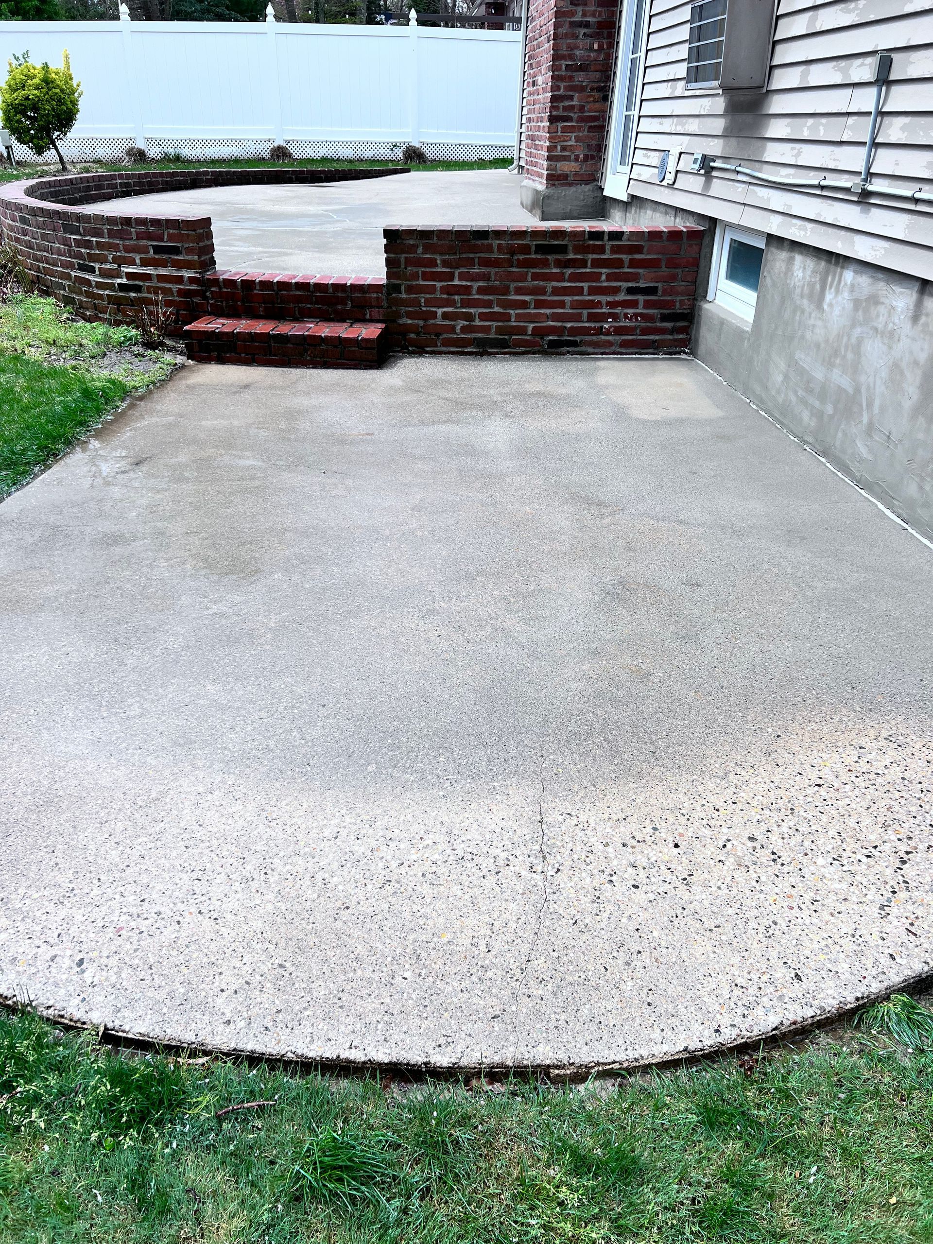Concrete patio bordered by grass and brick. A small brick wall and steps lead to a higher level. A chimney and house siding are to the right.