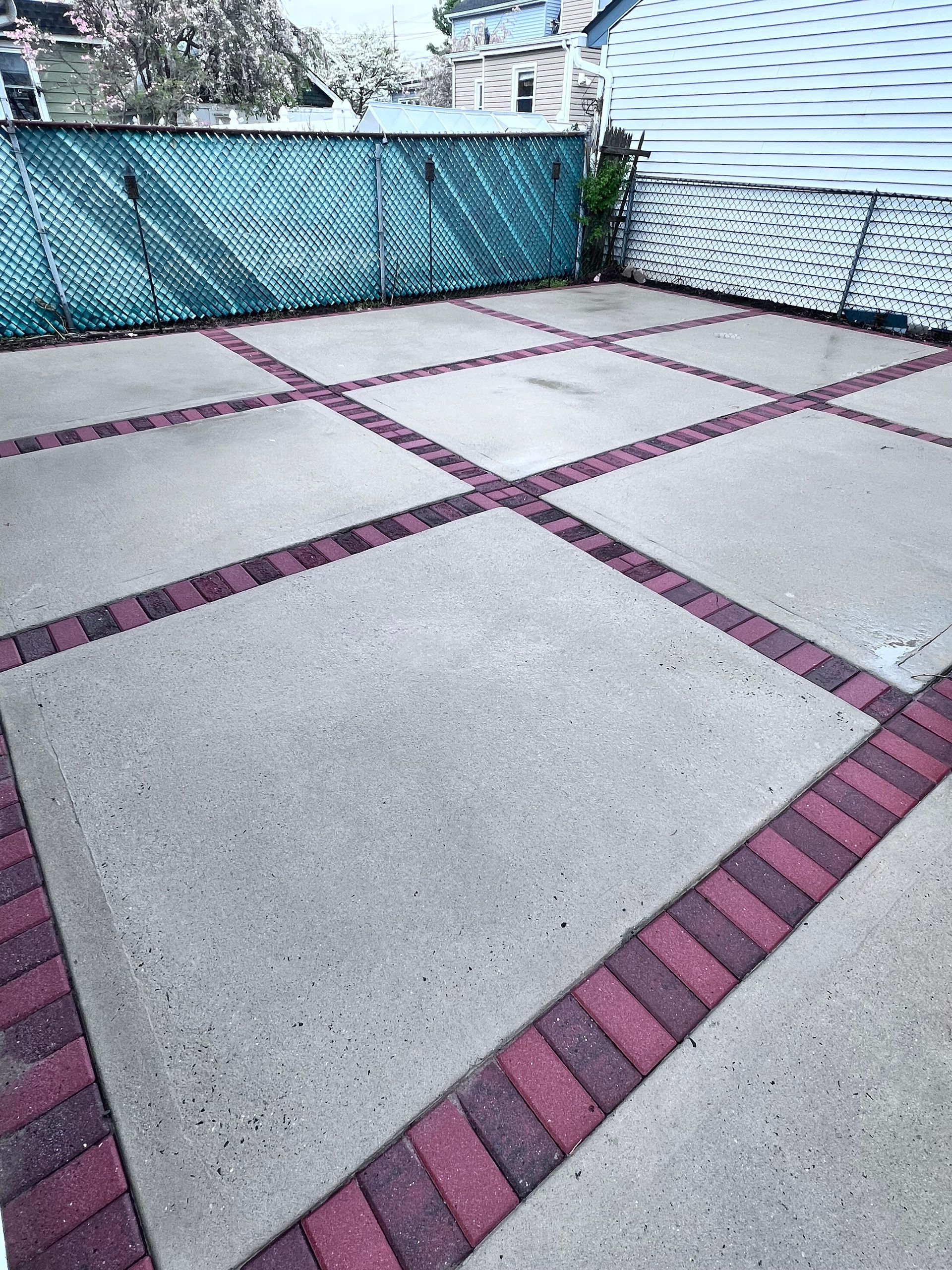 Concrete patio with brick border in a grid pattern, with a green privacy screen and a white-sided building in the background.