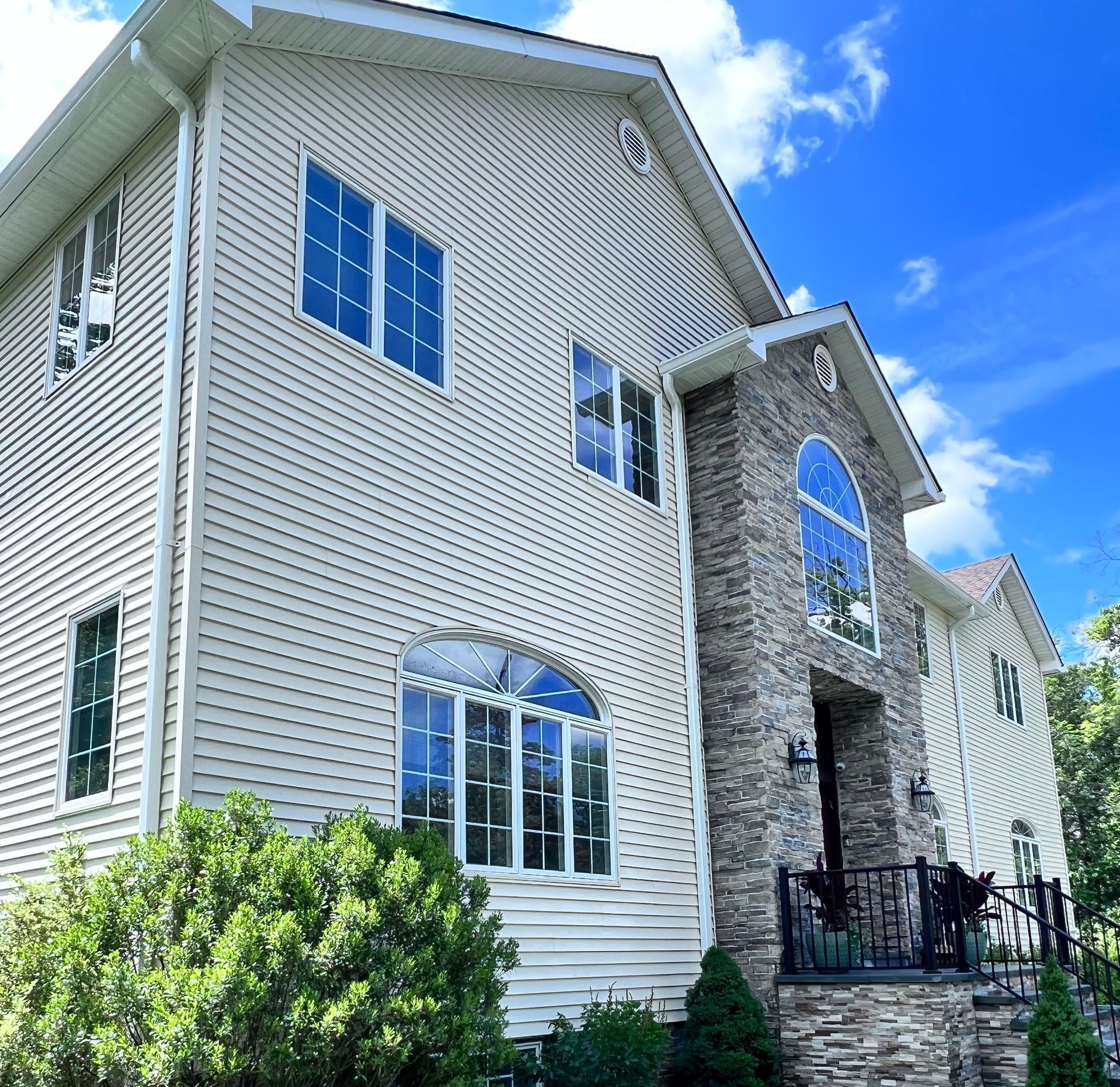 Two-story beige house with stone facade, blue sky background. Features arched windows and manicured landscaping.