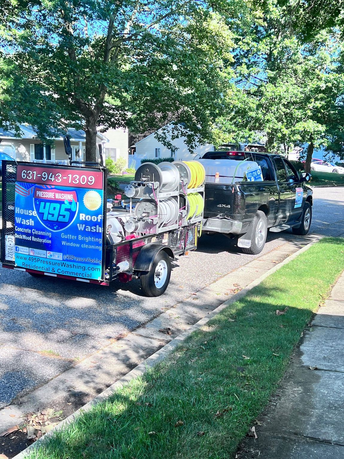 A black pickup truck towing a trailer with pressure washing equipment. The trailer has a large advertisement and is parked on a street with grass and trees.