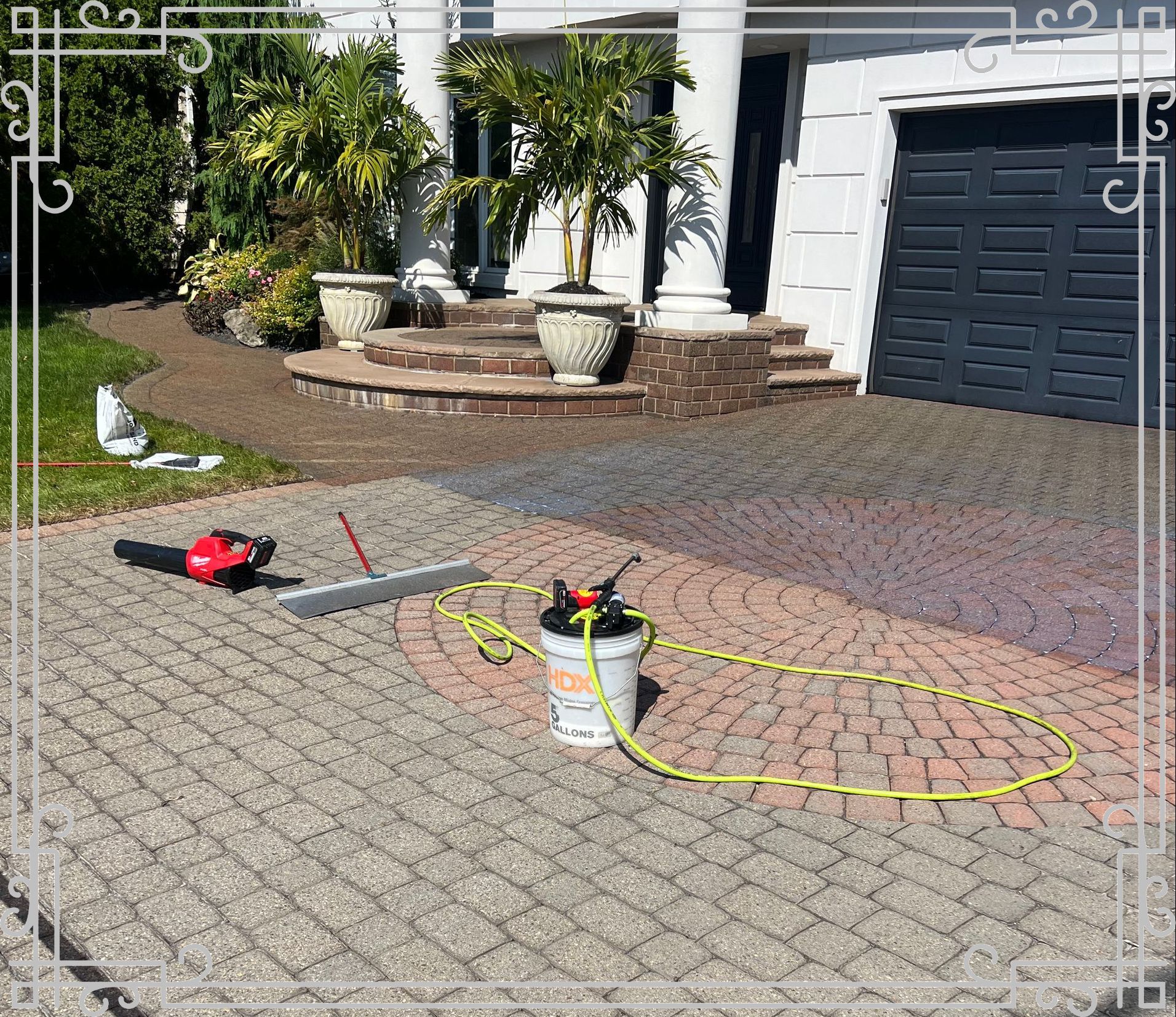 Driveway being worked on; tools like a sprayer, saw, and leveler sit on the brick pavers. A house with pillars and a garage door is in the background.