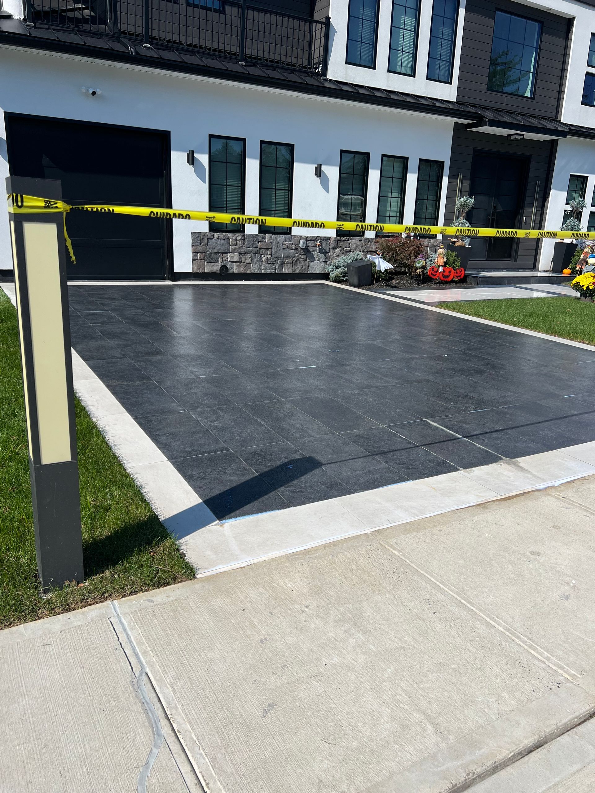 Driveway blocked by caution tape in front of a modern house with a black garage door and dark gray driveway. A light post is on the left.