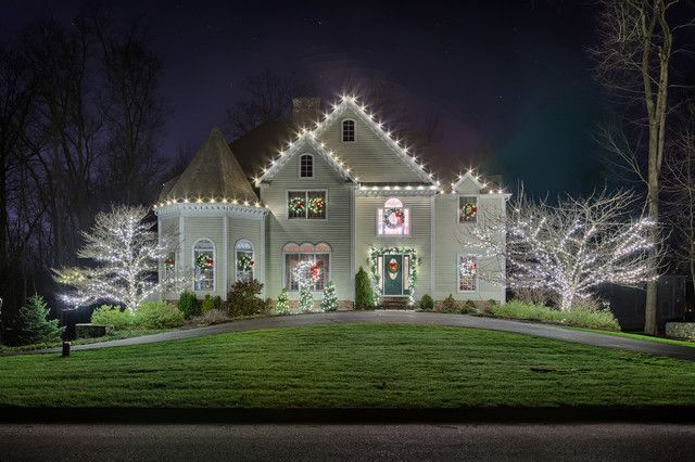 A house lit with white Christmas lights at night. Lights outline the roof, windows, and trees in front.
