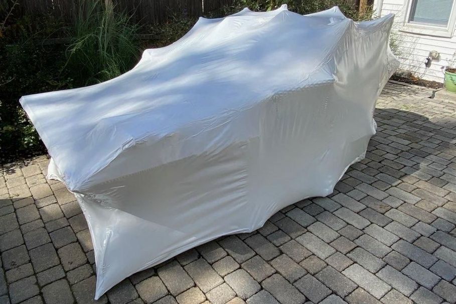 White plastic-wrapped object on a brick patio outside, possibly protecting furniture. Bright sunlight and greenery are visible.