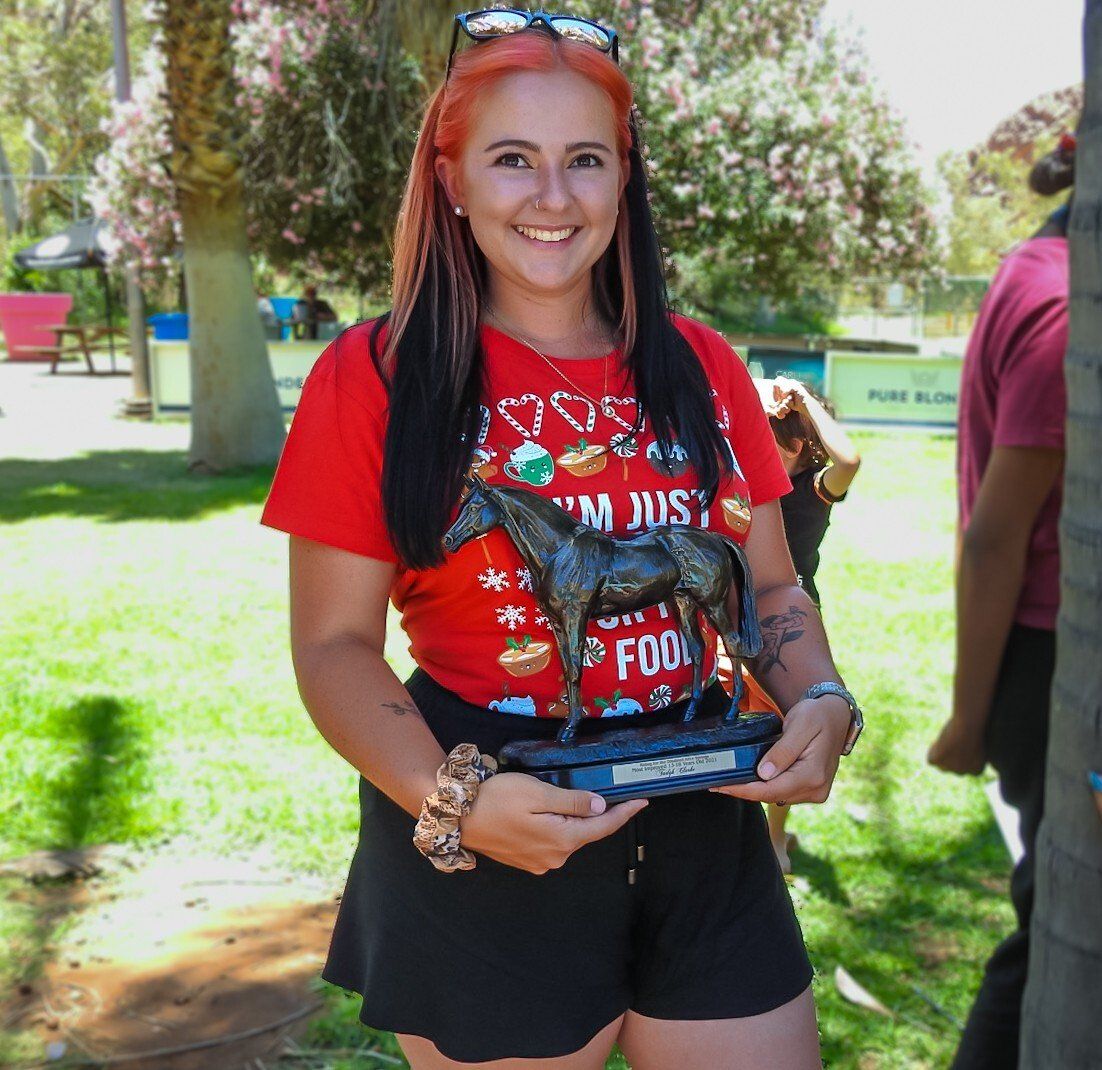 Woman with red hair and black shorts holding a trophy, smiling outdoors. 