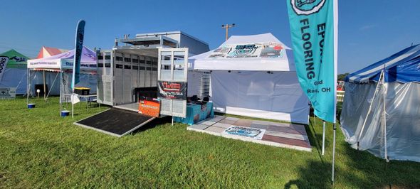 Outdoor event booth with white tent, teal flags, grass, and a small ramp display under blue sky