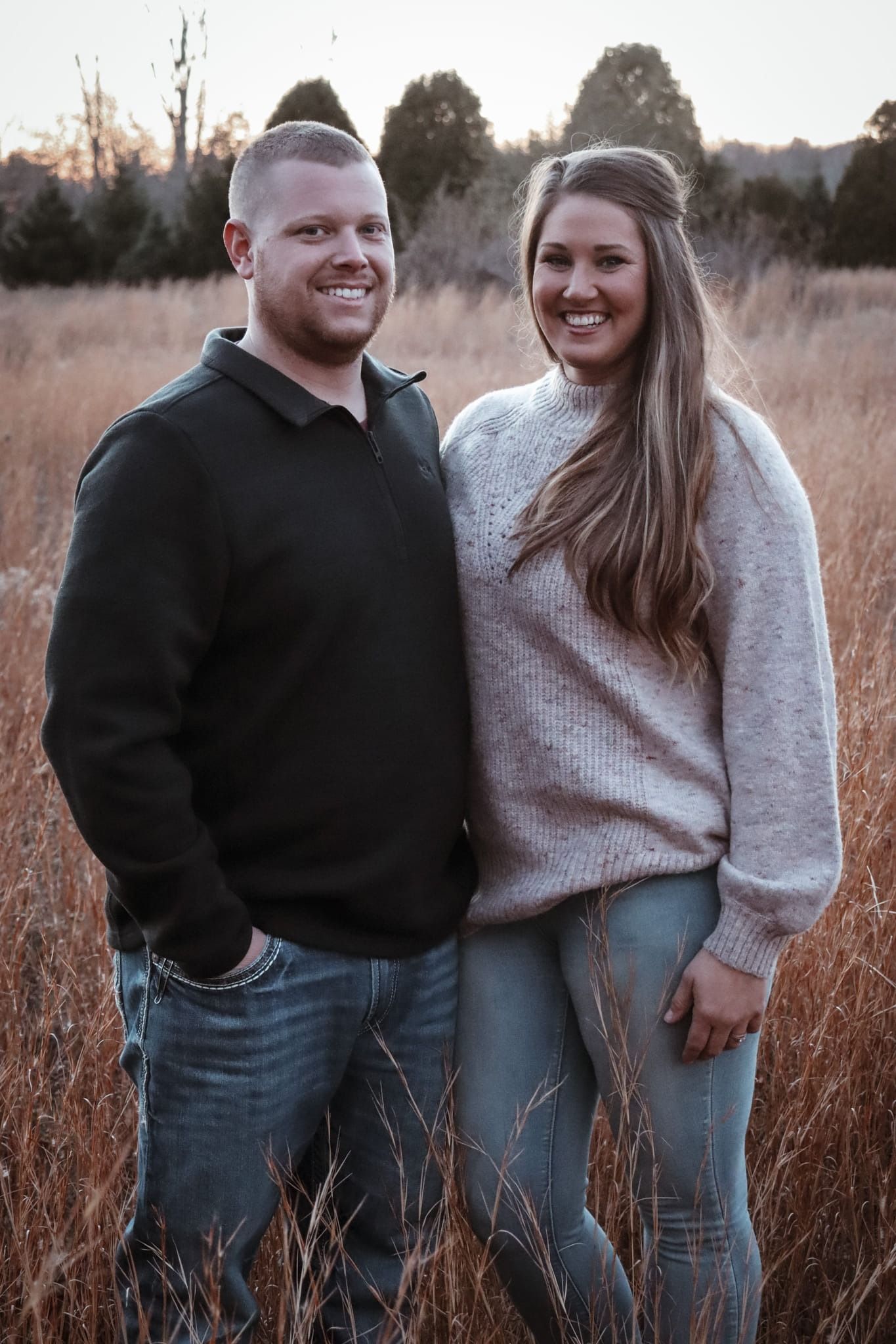 Two people standing in tall grass at sunset, smiling at the camera.
