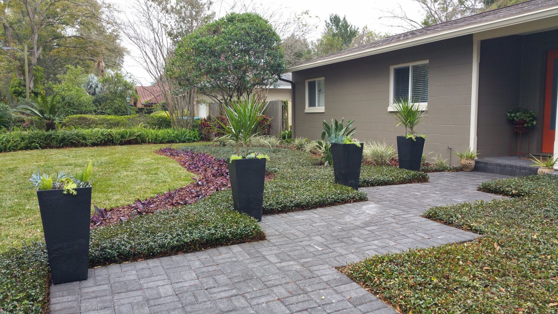 Front yard landscaping with brick pathway, rectangular planters, and green and purple plants.