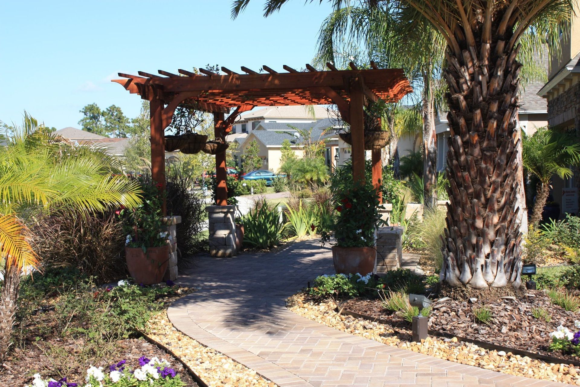 Arched wooden pergola over a brick path in a landscaped garden with potted plants and palm trees.